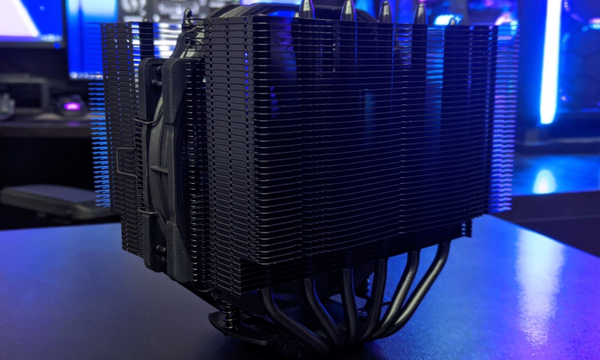 Close-up of a large black CPU air cooler with stacked metal fins and heat pipes, illuminated with blue lighting, dark moody tech desk background