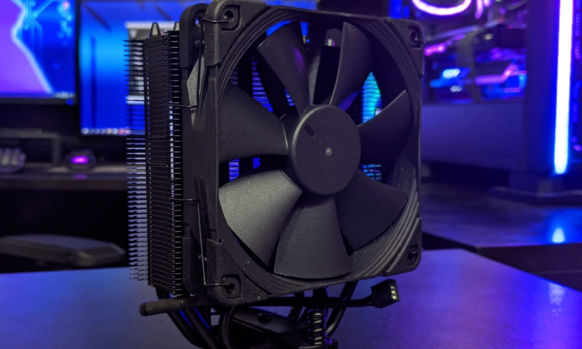 Close-up of a black cooling fan mounted on a heatsink with heat pipes, purple and blue ambient lighting, tech desk background